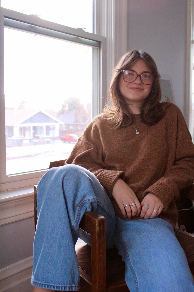 woman with glasses sitting on a chair in front of a window with her leg over the arm of the chair
