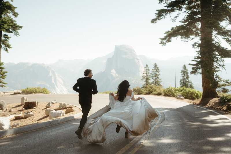 Bride and Groom run on the road during their Yosemite Elopement with bride's dress flowing behind her