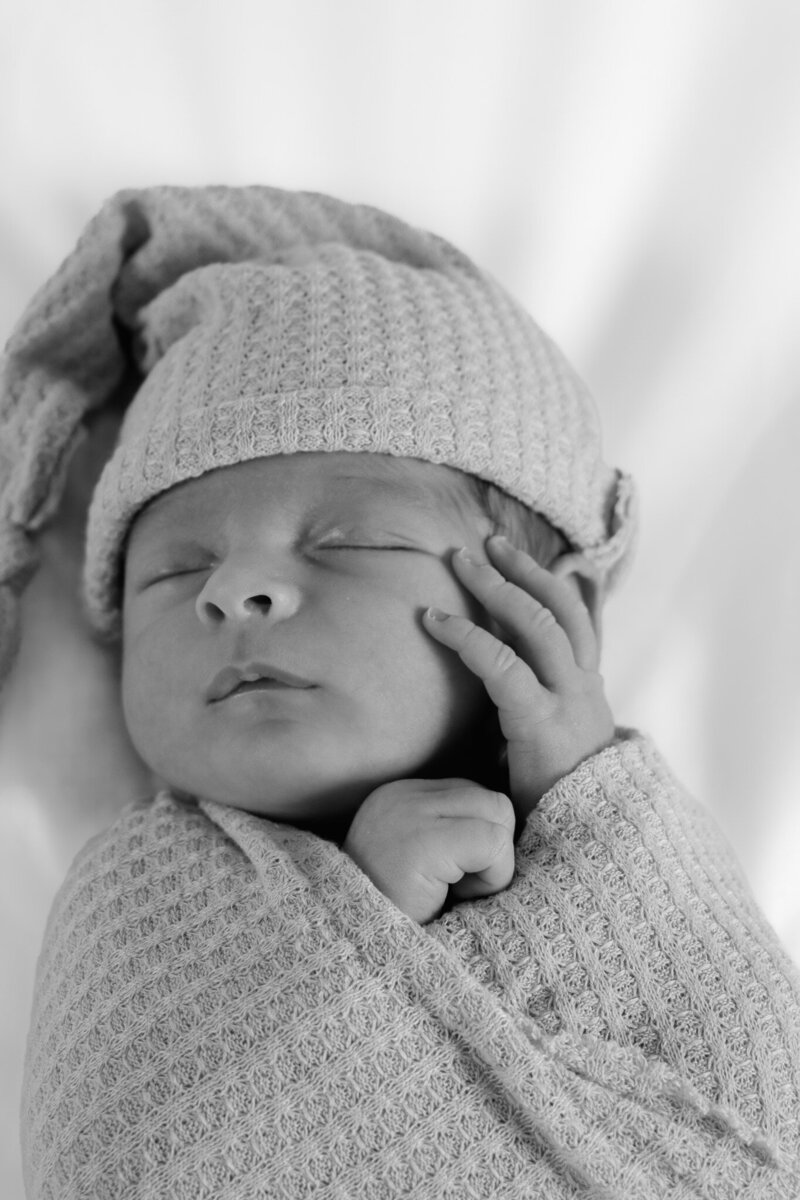 Black and white Newborn in a soft pose with arms tucked under the head, wearing a neutral-toned bonnet. Photographed with gentle elegance by Sophie Louise.