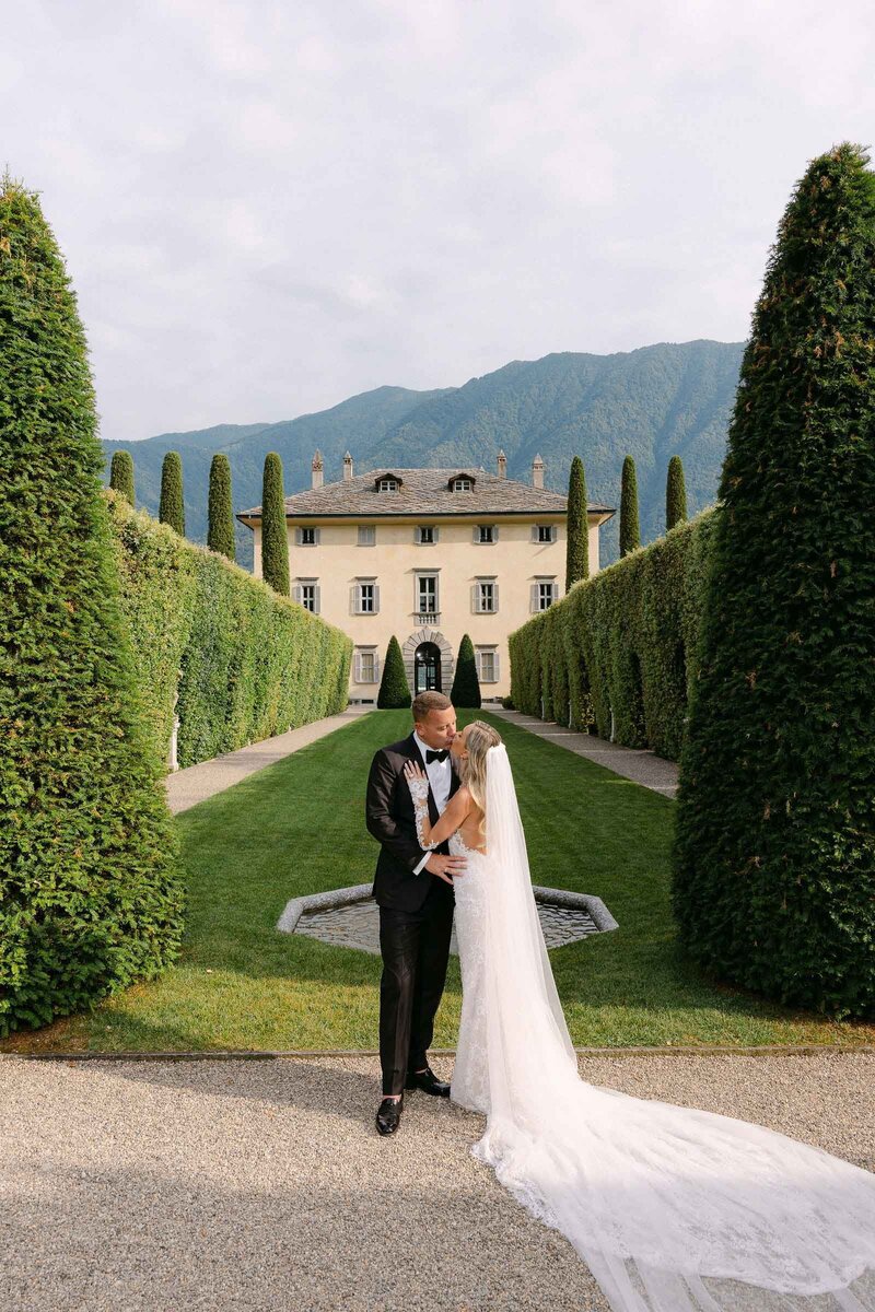 Bride and groom kissing at Lake Como with Villa Balbiano in the background.