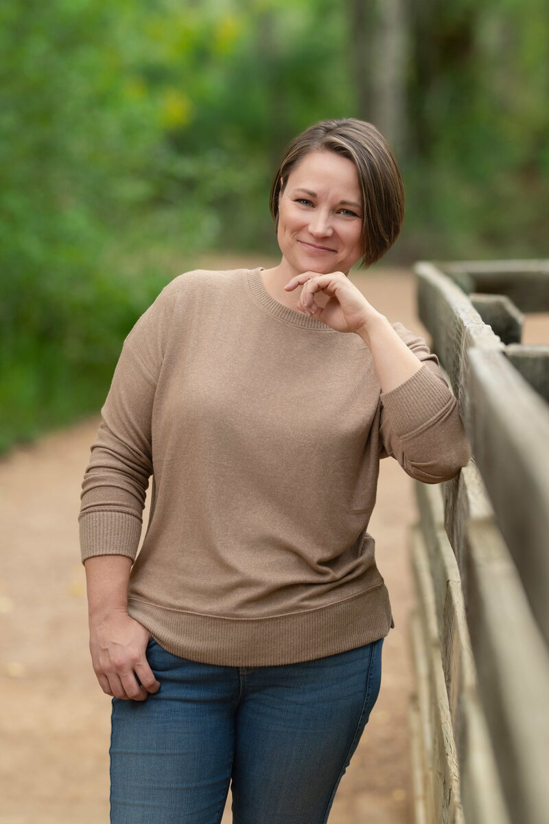 woman in brown sweater leaning on a fence in a wooded area