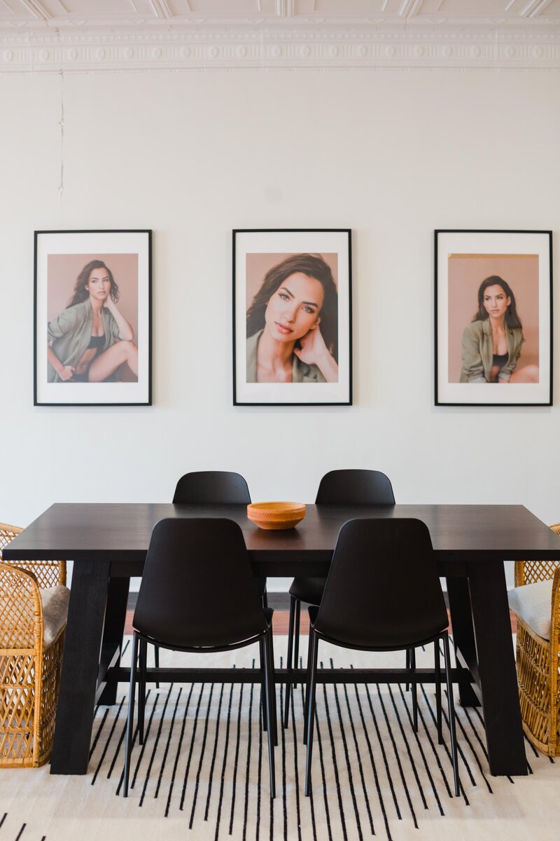 Black wooden table surrounded by four modern black chairs sits on a black and white rug in front of a wall with three framed portraits of a woman | Sara Coffin Photo