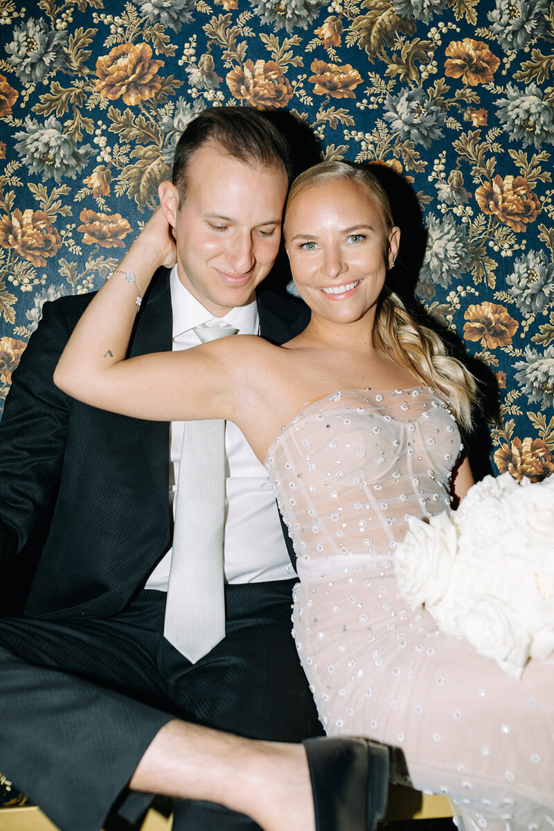 Blonde bride in chic bridal gown smiles at camera and wraps her hand around her groom's neck.