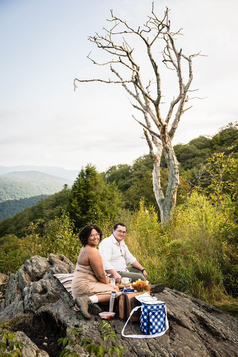 A couple on their elopement day in Shenandoah National Park sit on a large rock at Jewell Hollow Overlook for their breakfast picnic.