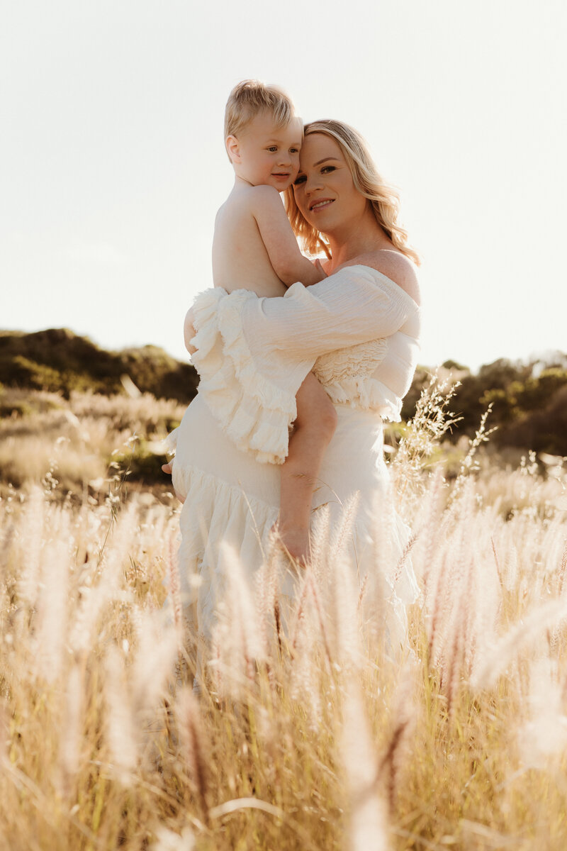 A family of four stands by a riverbank at sunset. The father holds a baby, the mother holds the hand of their young child. Dressed in casual summer outfits, they enjoy the serene backdrop of trees and water, captured beautifully by their trusted Illawarra newborn photographer.