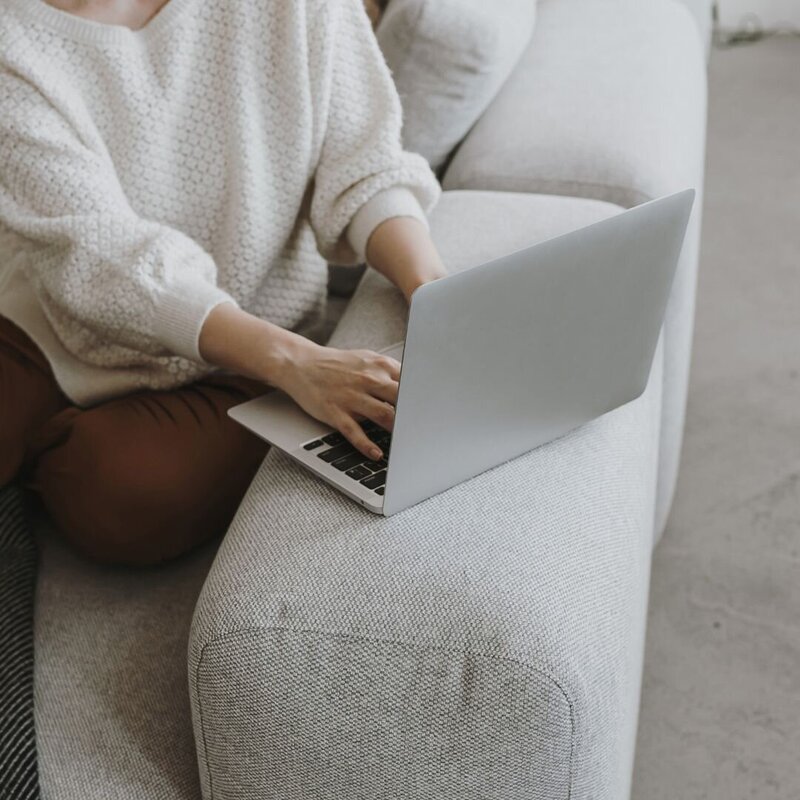 Someone typing on their laptop while sitting on a couch and wearing a white sweater and brown pants.