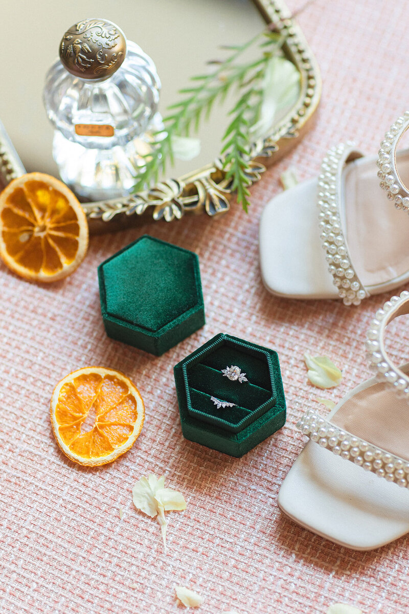 Flat lay photo of shoes, orange slices, and wedding rings in a dark green ring box