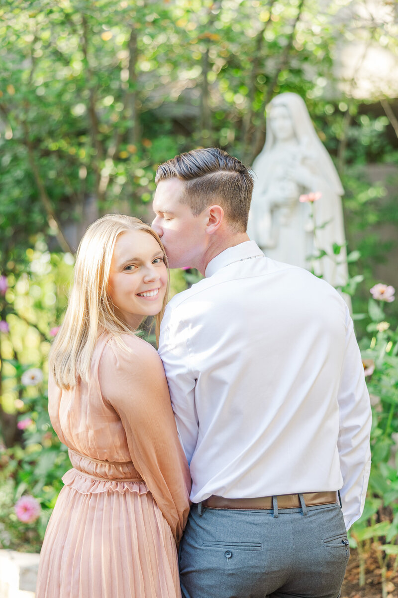 a man kissing his fiance on the head while they stand in front of a Catholic statue