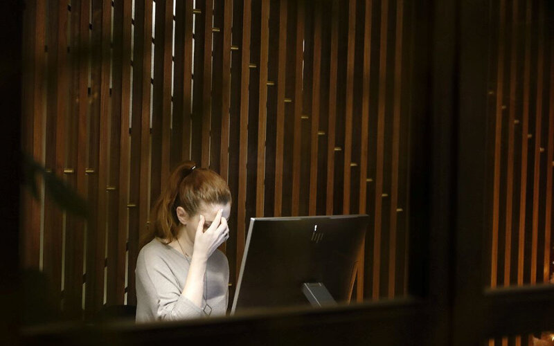 An overwhelmed young woman sits at her desk, stroking her forehead like she has a headache.