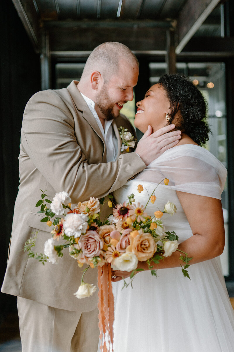 A bride and groom laughs together and holds each other close while holding an Anthousai bouquet.