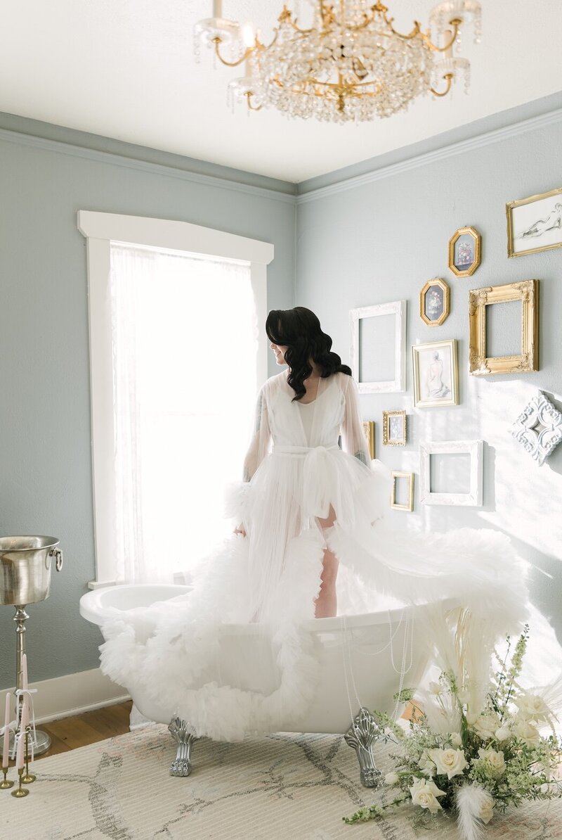 Editorial bridal portrait of bride standing in a clawfoot tub wearing a dramatic tulle robe inside a vintage-inspired bridal suite at The Manor House in Colorado