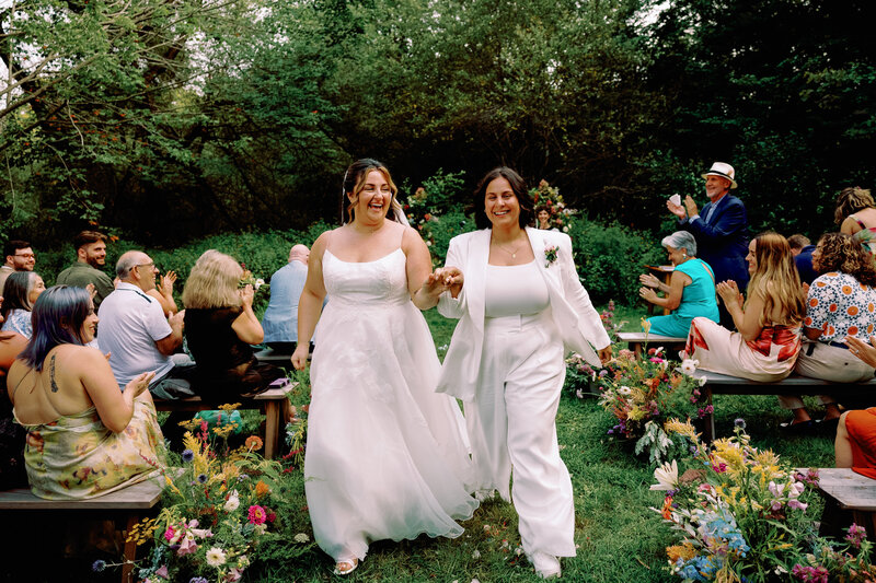 two brides walking down the aisle after getting married with bright florals outside