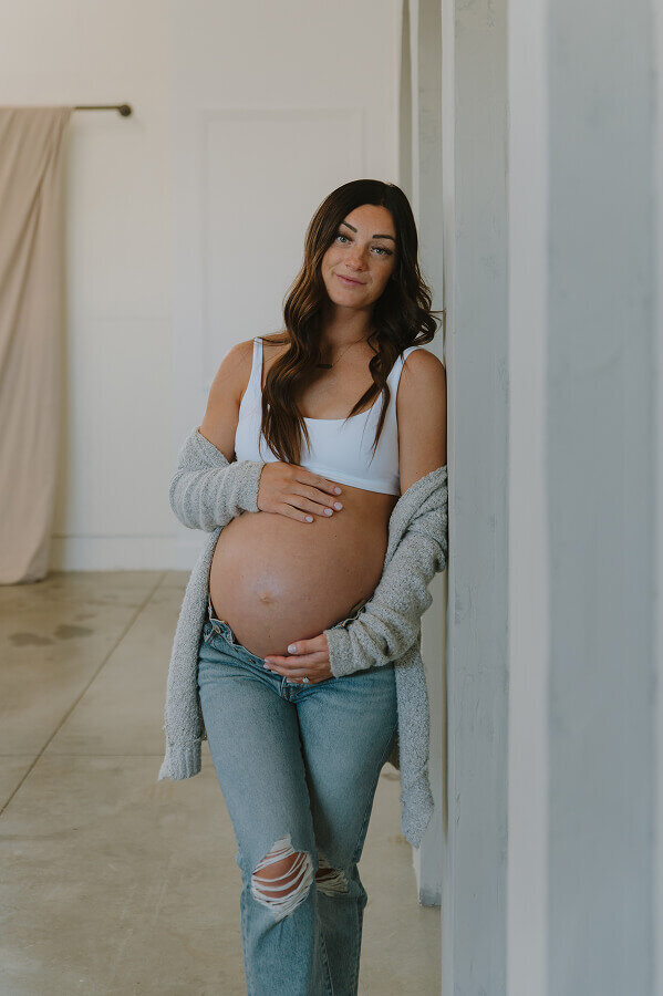Wearing a white sports bra and jeans, a woman lovingly holds her pregnant belly. She's standing inside of a photography studio.