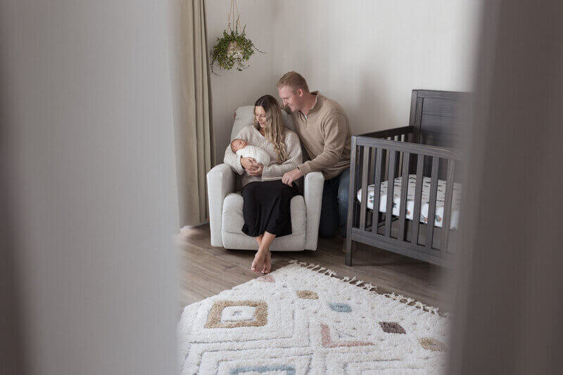 A cozy family moment in a softly lit nursery, with a mother cradling her newborn while sitting in a beige armchair, and the father leaning in lovingly beside her, next to a dark wooden crib.