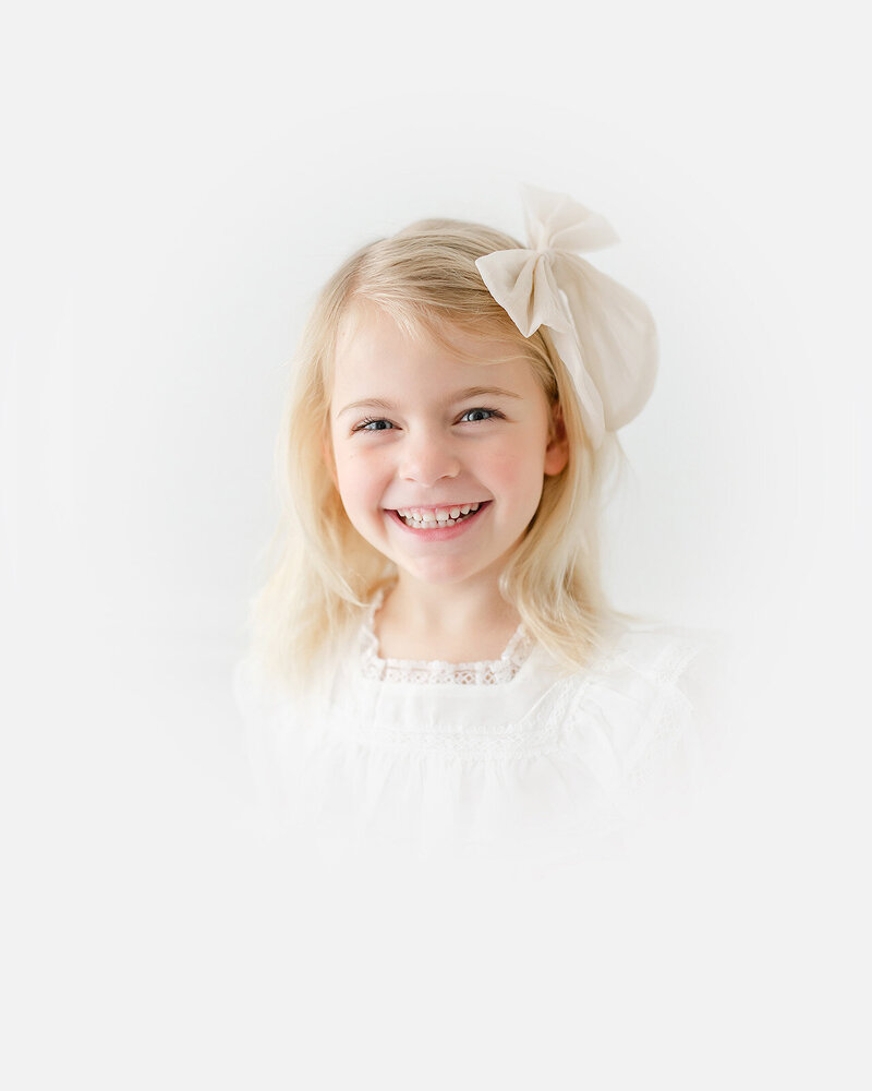 A smiling young girl with blonde hair and a large bow, wearing a white dress in an heirloom portrait, stands against a light background.