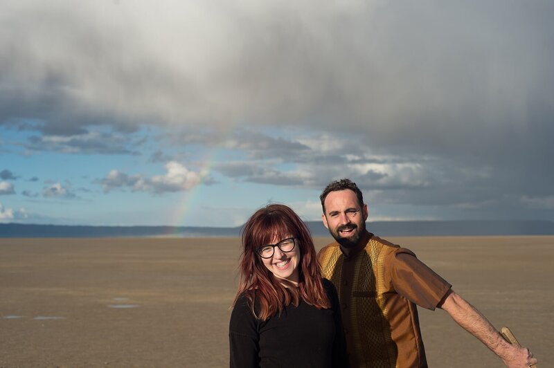 a couple stand in the middle of the alvord desert with a rainbow in the background