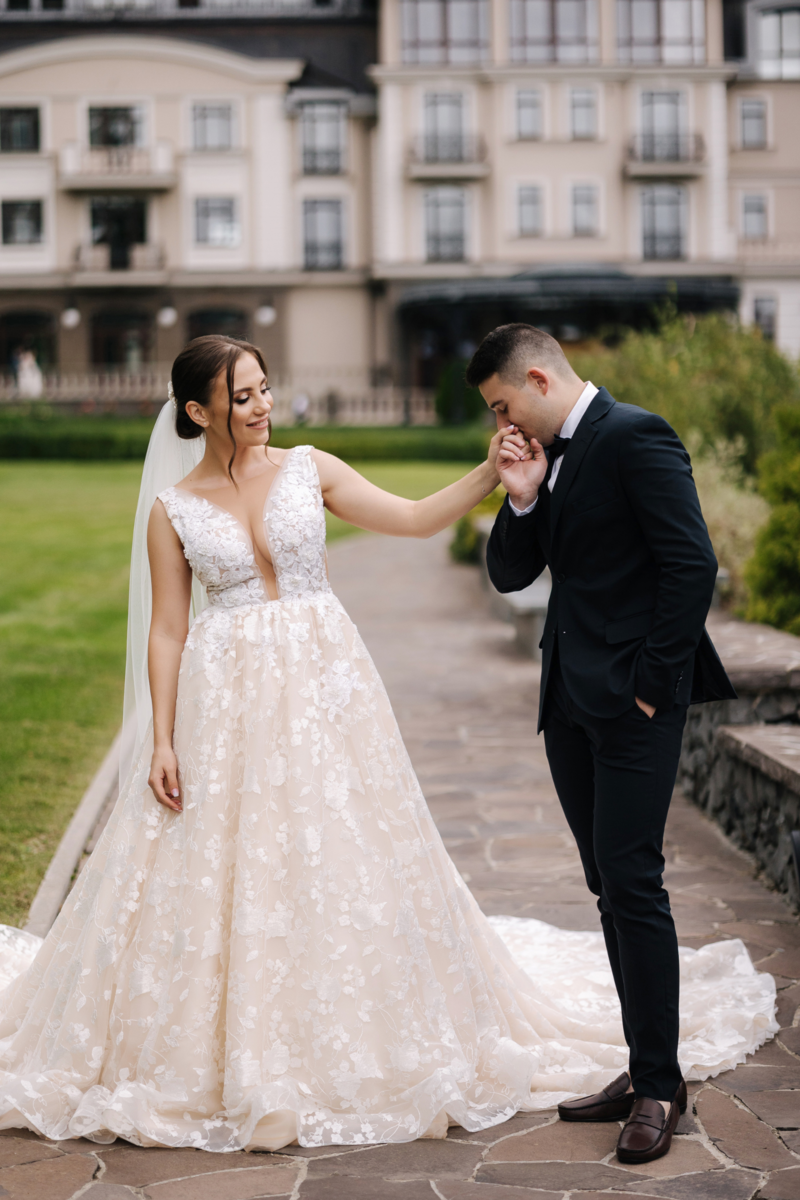 A tender moment captured as the groom kisses the bride's hand outside the wedding venue, symbolizing love, respect, and affection in a romantic gesture.