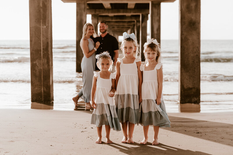 Three young sisters smiling with parents in the distance smiling while all standing under a pier in Port Aransas, TX.
