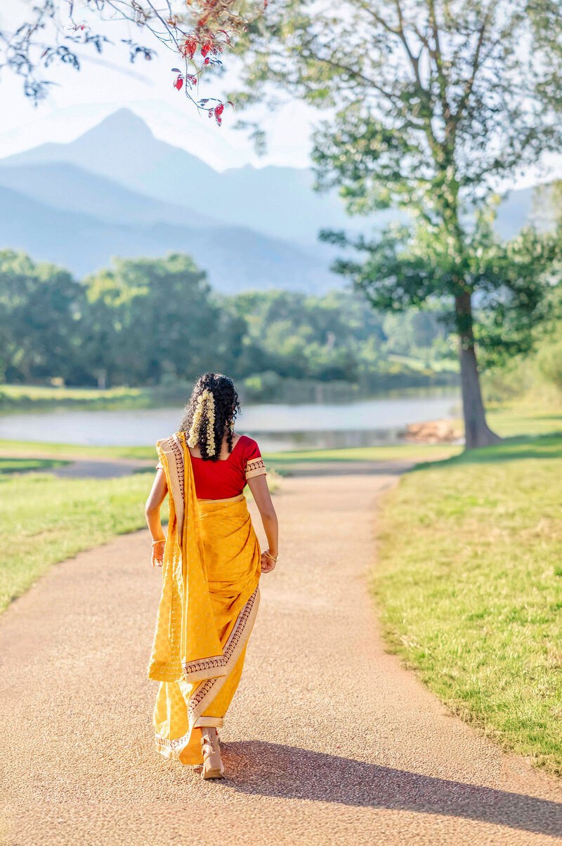 A woman in a yellow sari and red blouse walks down a path surrounded by green grass and trees. A lake and mountains are visible in the background under a clear blue sky.
