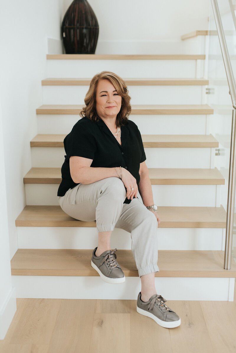 Joanne McCrone, Port Moody real estate advisor, seated confidently on modern wood stairs in a bright home.