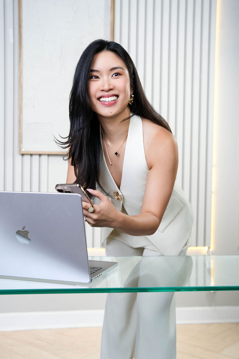 Stephy Chen smiling while working on her laptop at a glass desk, captured during a lifestyle personal branding photoshoot.