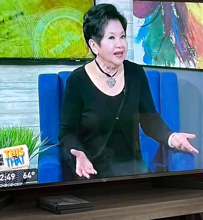 Nita Tin speaking with animated gestures during her interview on Channel 9 Chattanooga’s This and That. She is seated in a blue studio chair, wearing a black shirt and sweater, sharing her story with host James Howard.