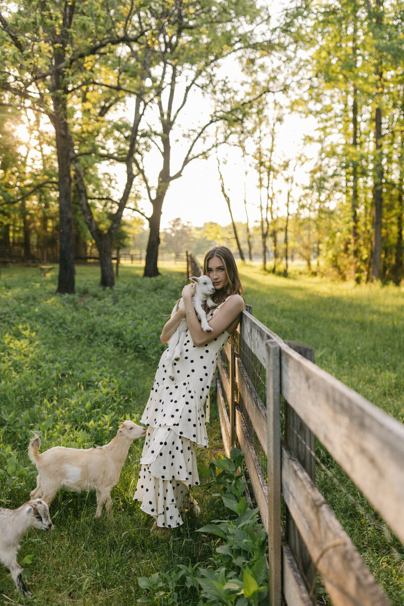 Sun-drenched portrait of a woman in a polka dot dress holding a baby goat on a wooden fence in Greenville, South Carolina. Captured by Dani Lacey Photographs.