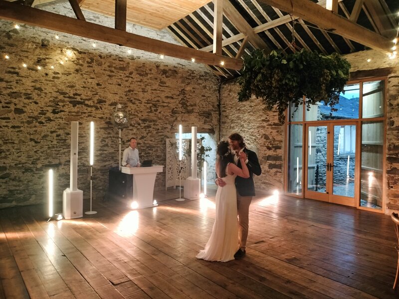 Bride and groom sharing their first dance in a rustic indoor wedding venue with exposed stone walls, wooden floors, ambient lighting, and a DJ in the background.