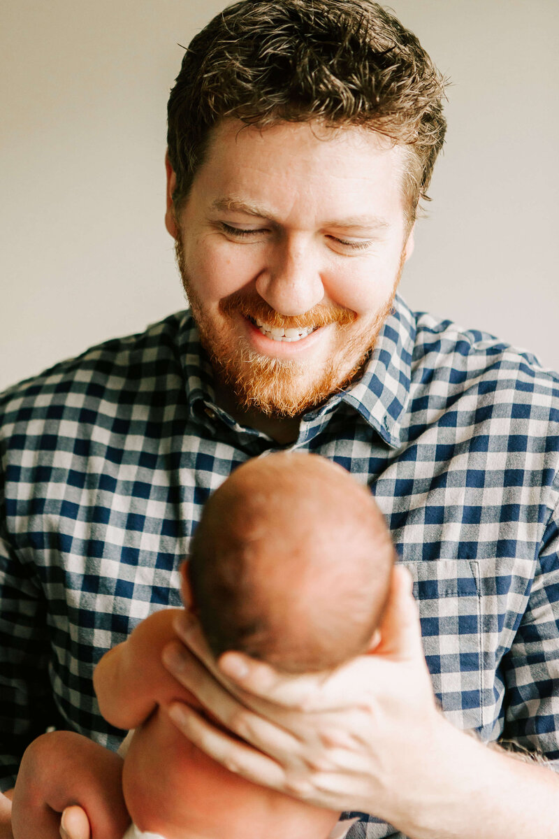 A dad in a plaid shirt is smiling and holding his newborn baby