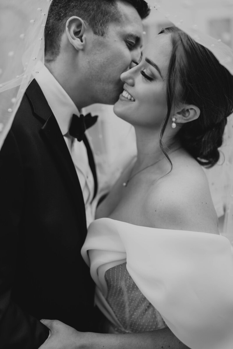 Black and white portrait of groom kissing bride on the cheek.