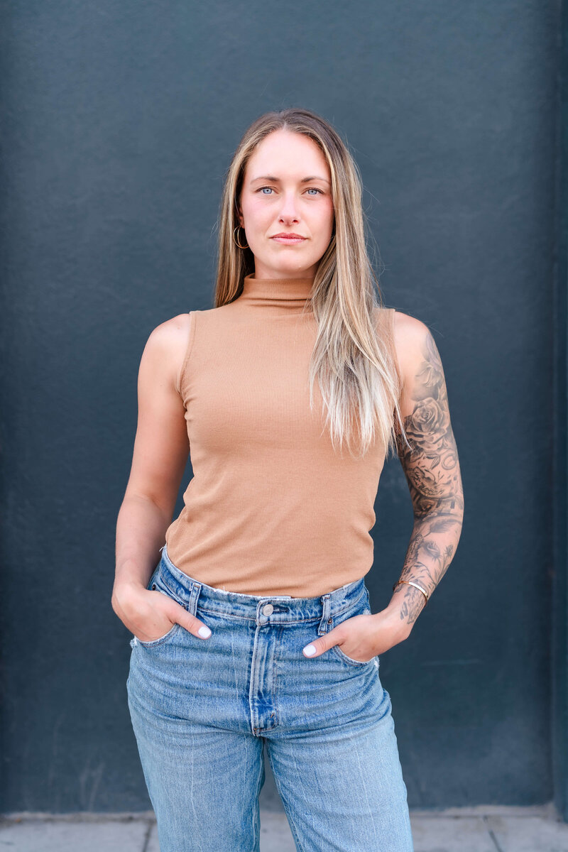 Strong woman in sleeveless tan top and ripped blue jeans posing confidently against a dark gray background for casual headshot.
