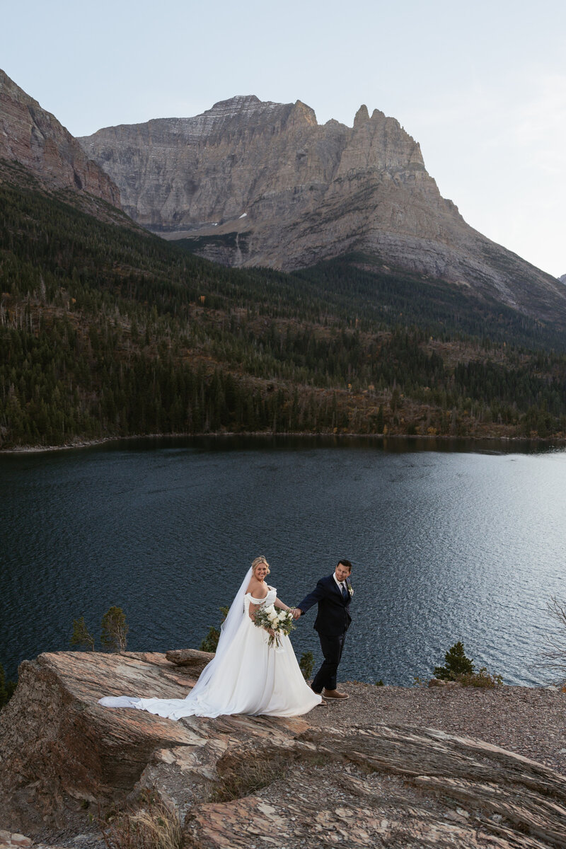 A couple holds hands and walks on the shore of an alpine lake in Glacier National Park.