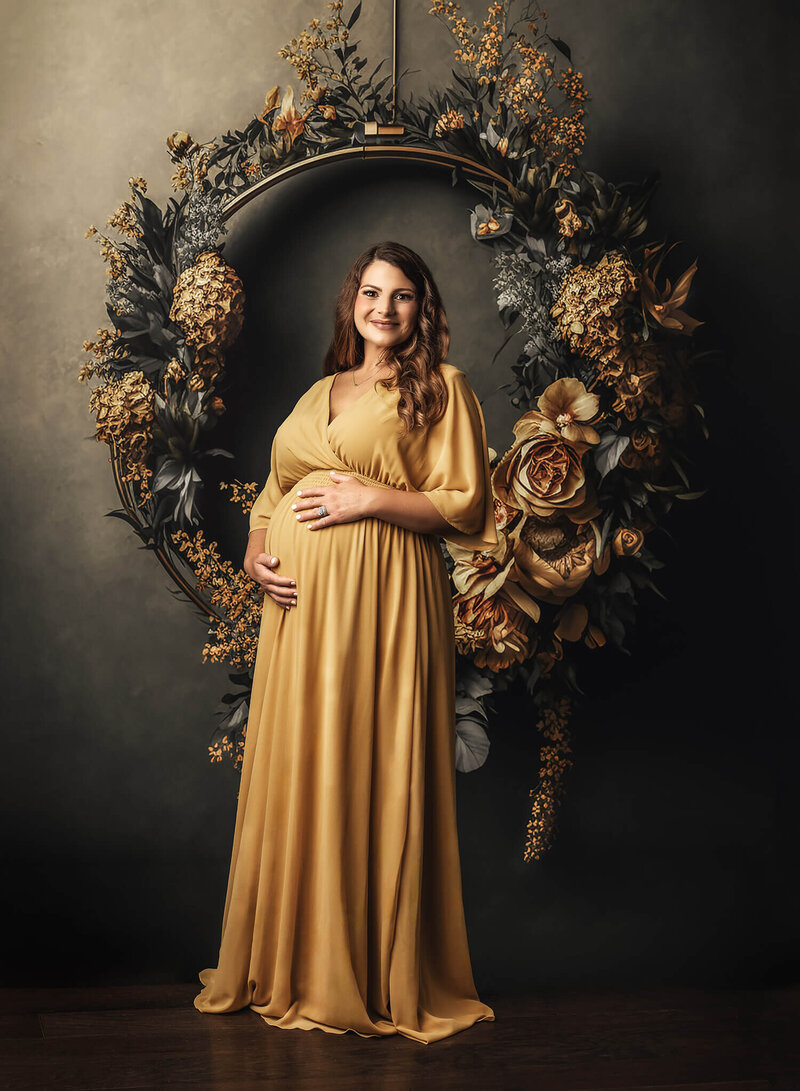 mother wearing golden dress with hands placed on her pregnant belly in front of a golden wreath of flowers