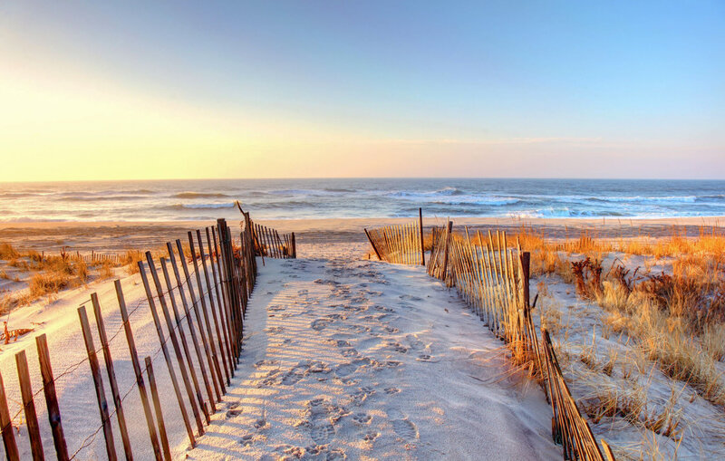 A sandy path with footprints leads to a serene beach. Wooden fences line the pathway, and dried grass is visible on the sides. The ocean waves are gently crashing, and the sky is a gradient of soft oranges and blues at sunset.