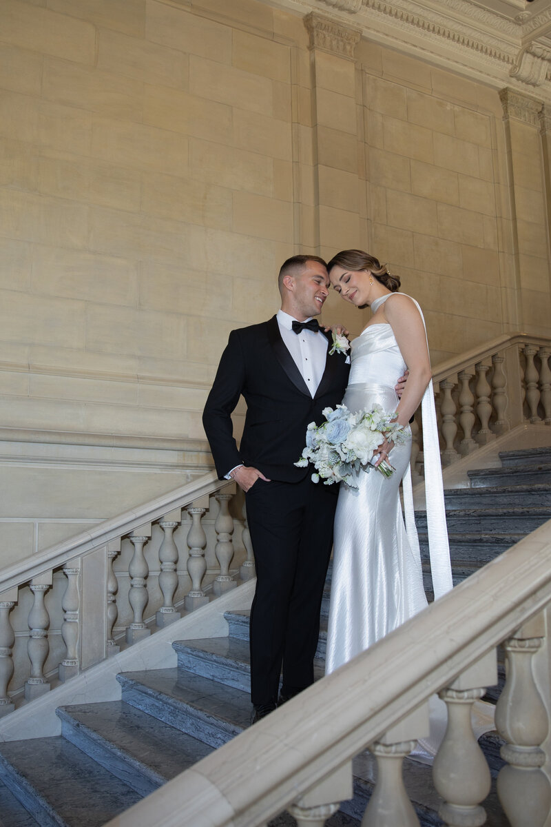 Rhode Island Wedding Photographer | Kelsey Sheehan Photography Timeless Rhode Island Weddings | A bride and groom pose on an elegant, curved staircase. The bride, in a satin gown, holds a bouquet; the groom wears a black suit and bow tie. Both look lovingly at each other.