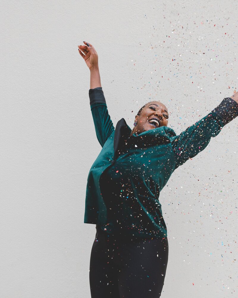 Black woman in a dark turquoise velvet suit, smiling, arms overhead, confetti falling around her. Photo by Clay Banks via Unsplash.
