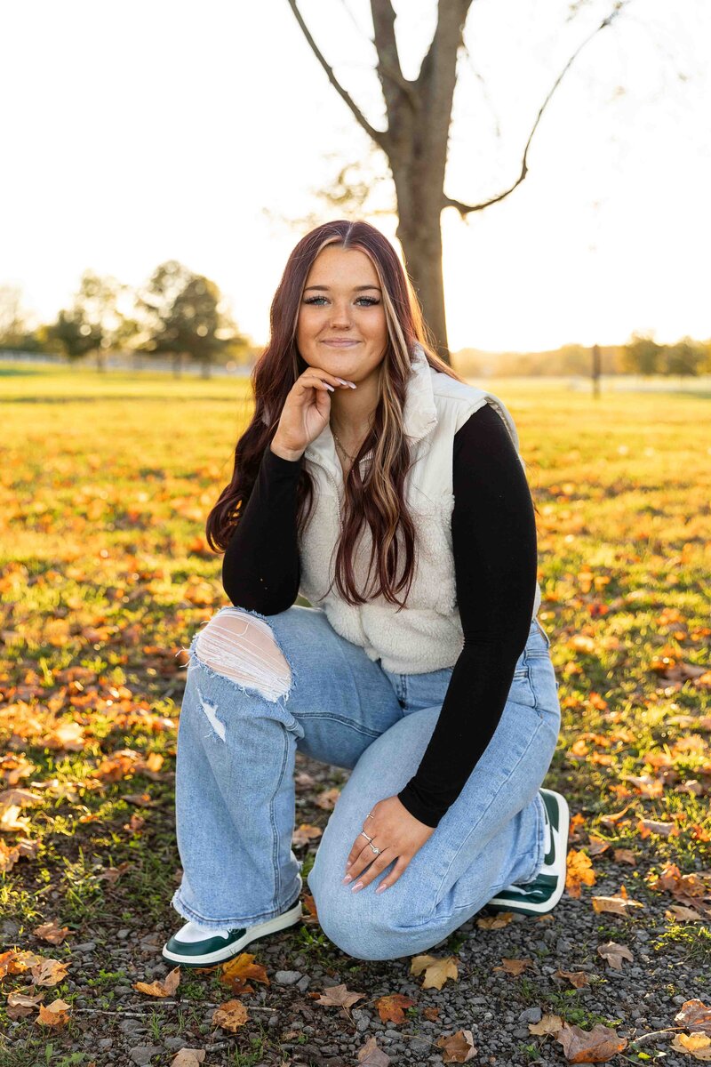 High school senior girl kneeling on ground with hand under chin smiling in the sunlight