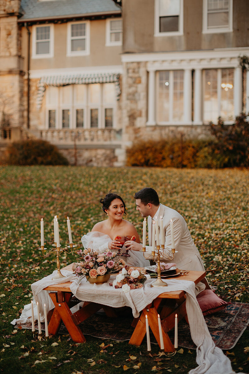 Elopement picnic: The newlyweds sit behind a beautifully adorned table on a lawn covered with golden autumn leaves.