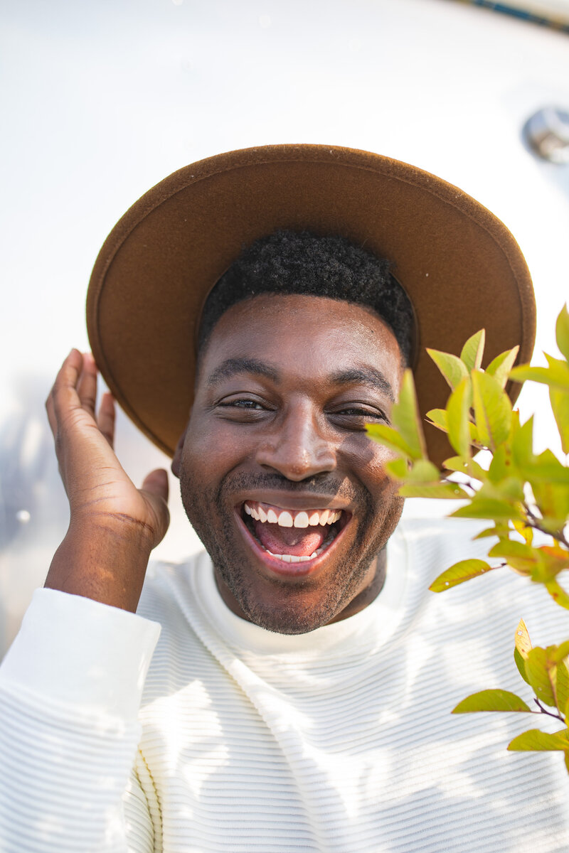 A man with a big smile, wearing a brown hat and a white sweater, is laughing joyfully. He is outdoors, with a plant partially visible in the foreground, and the background is bright and sunny.