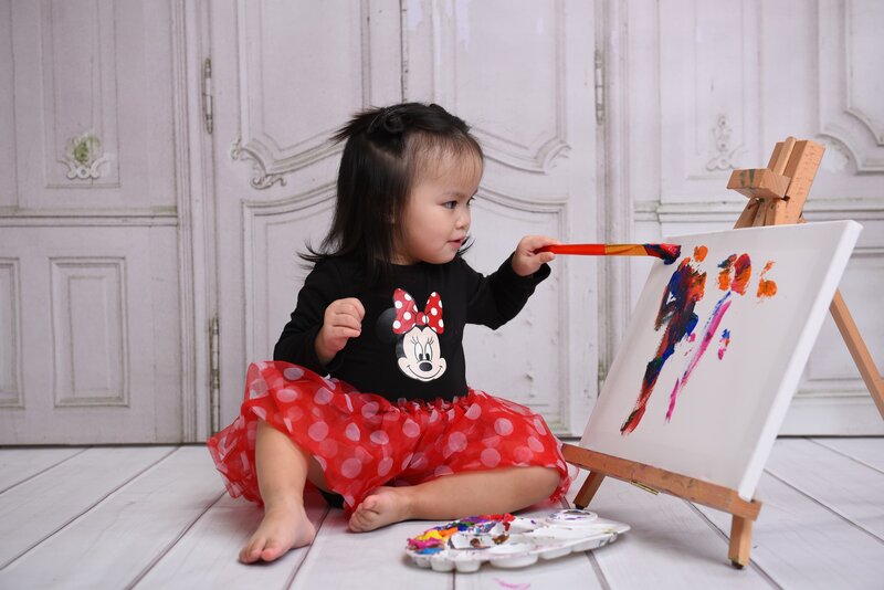 A toddler dressed in a Minnie Mouse-themed outfit painting on a canvas during a creative photo shoot. The background features elegant white doors, adding a playful and classy touch to the session.