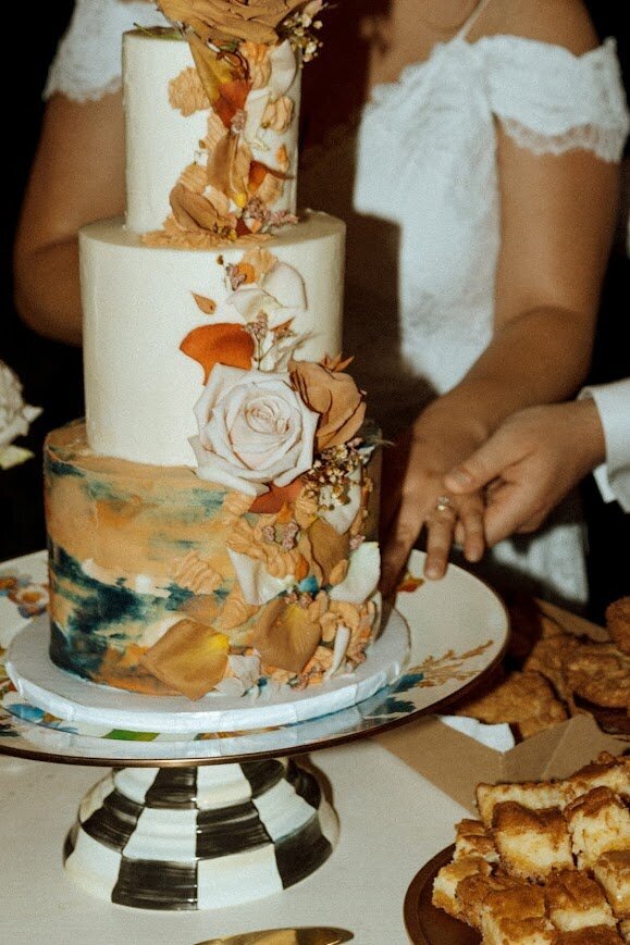 A couple cuts their wedding cake. Photo primarily just shows the brides torso and dress, with her hand being held by her husband as they cut the cake. The cake is a three tier wedding cake, with two white tiers on top, and a bottom tier with smudges in different shades of teal and terracotta. Down the side is a cascade of fresh roses, rose petals, and squiggles of frosting in the terracotta color.