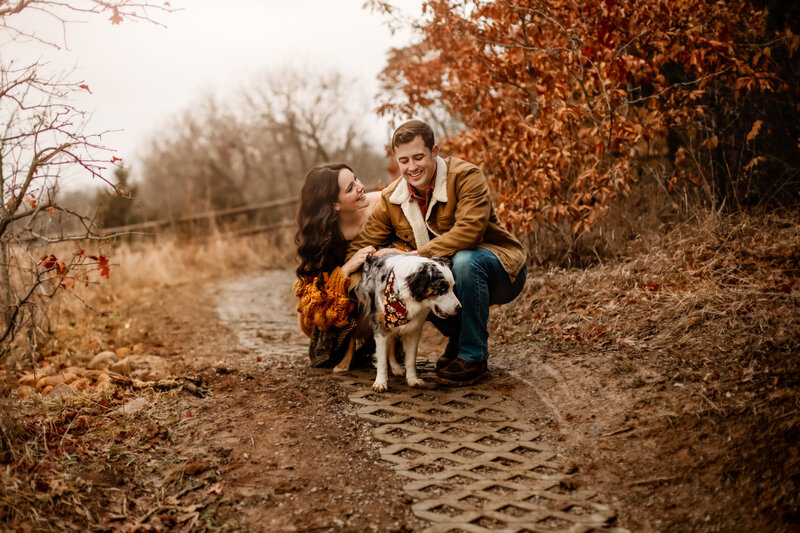 Husband and wife smile at each other and their dog on a walk way for a winter maternity session with Family Photography Kansas City photographers.