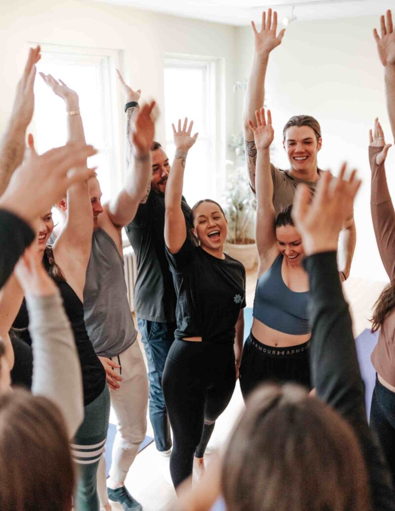 A group of breathwork participants raising their hands joyfully, celebrating connection and transformation in a light-filled studio.