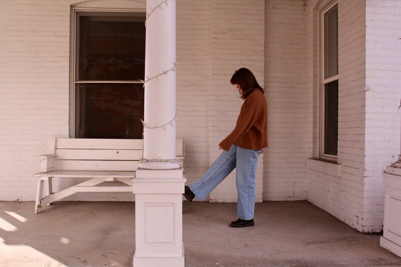 Woman wearing a brown sweater and jeans walking on a porch with white brick