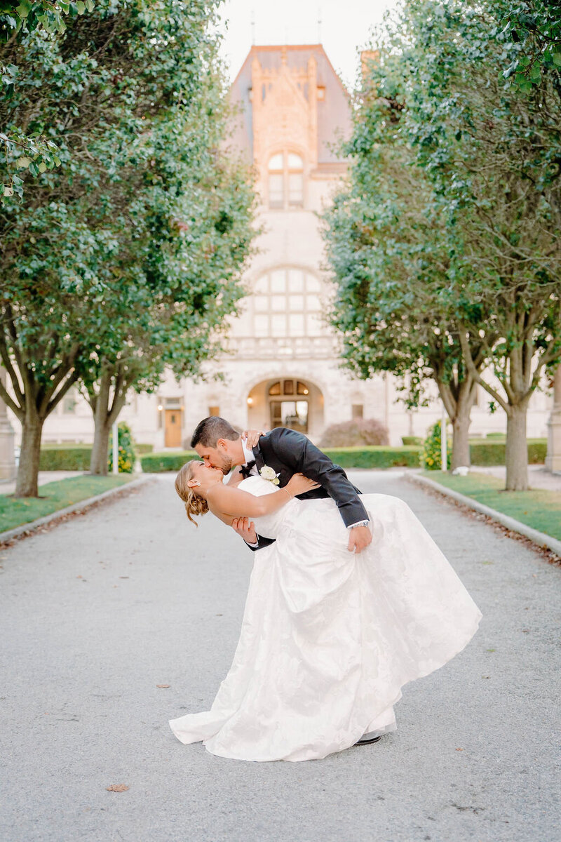 Rhode Island Wedding Photographer | A couple, dressed in wedding attire, shares a romantic dip kiss on a tree-lined path leading to a grand building.