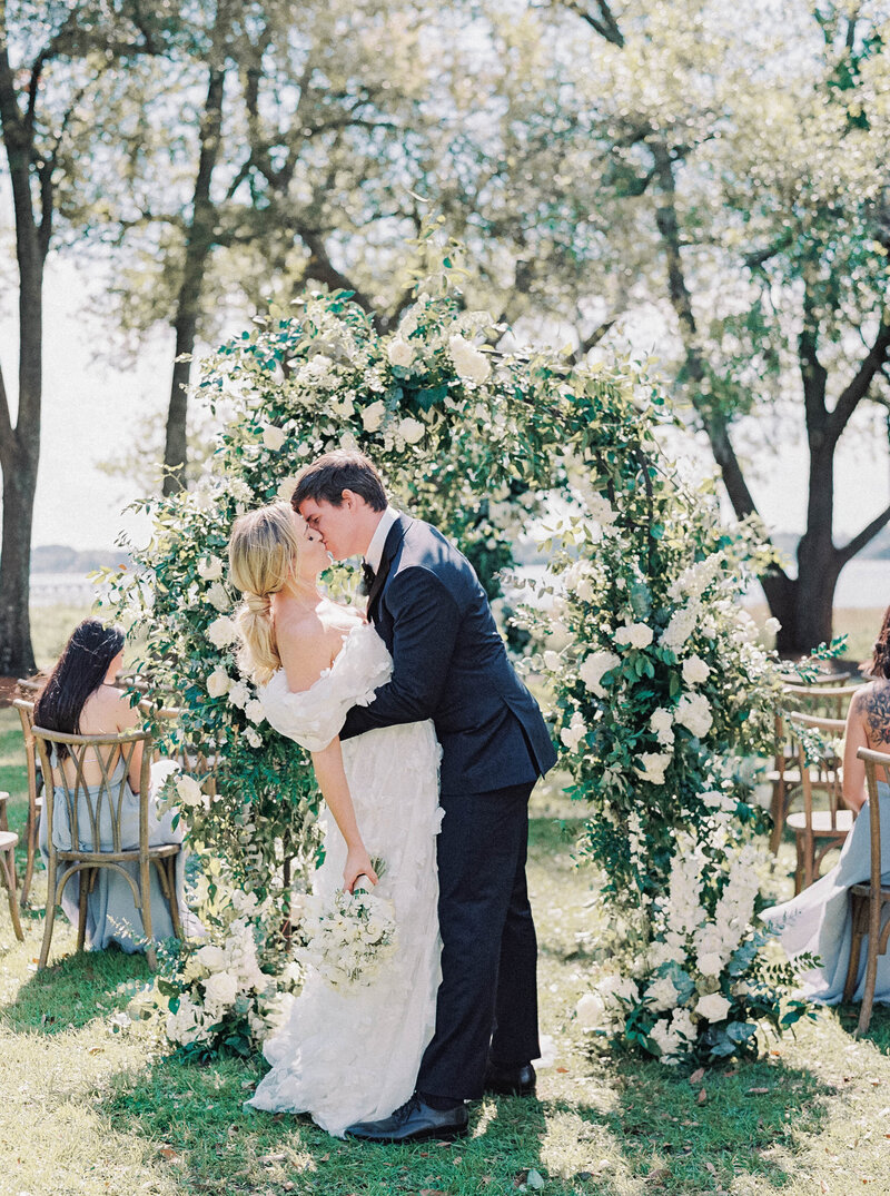 RI Wedding Photographers | A couple kisses under a lush floral arch adorned with white flowers and greenery at an outdoor wedding. Guests sit in the background under sunlit trees.