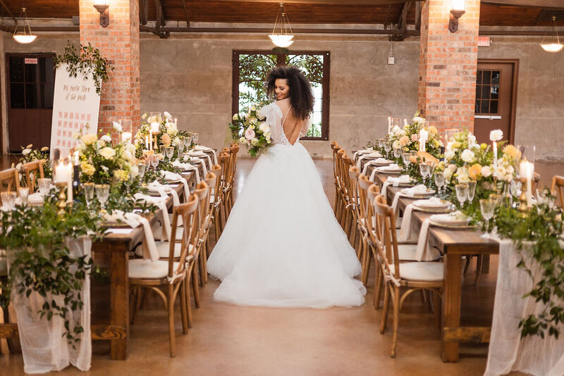 A bride stands between two rows of rectangular wooden farmhouse tables decorated with florals and crossback chairs in an industrial venue.