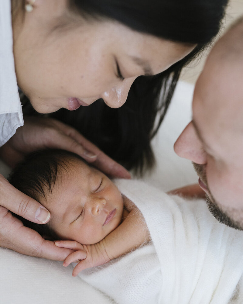 up close photo of a couple looking down at their asian newborn baby sleeping and swaddled in white lush angora rug, Perth newborn in home session