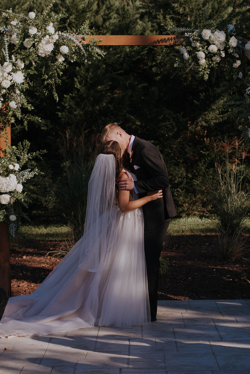 A couple in wedding attire shares a kiss under a wooden arch adorned with white flowers and greenery at a rustic wedding venue. The bride, in a flowing gown and veil, and the groom, in a black suit, pose on a stone path with lush Virginia greenery captured in the background.