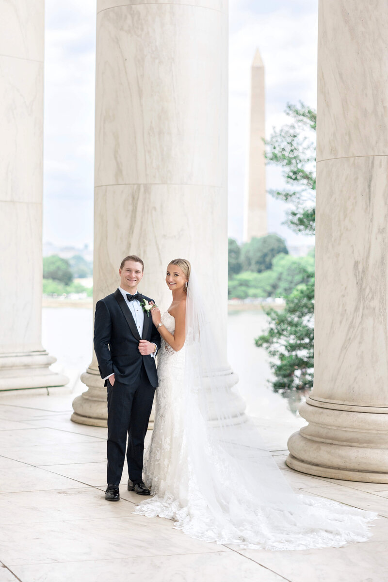 A bride and groom stand smiling in a marble columned area, with the Washington Monument visible in the background. The bride wears a long white gown and veil, while the groom is in a black suit and bow tie.